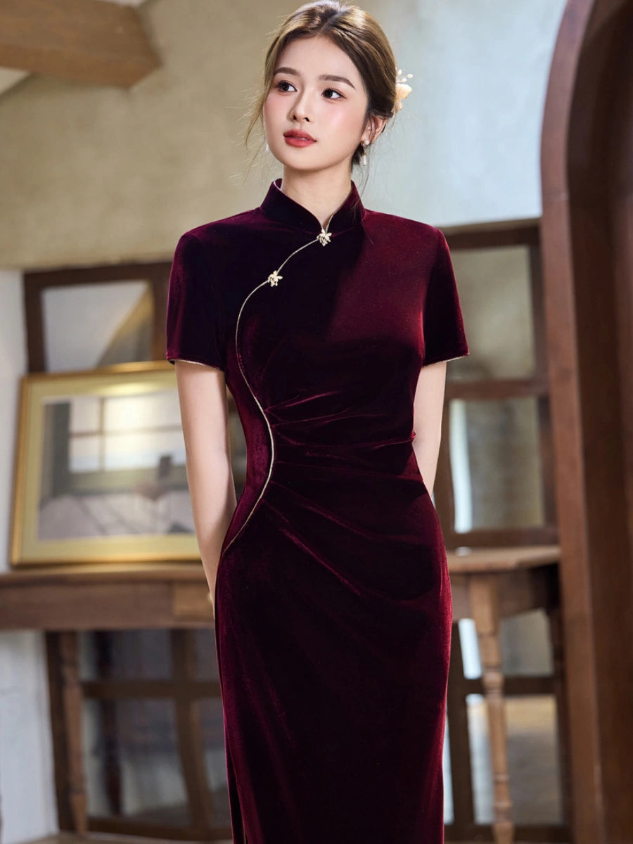 A woman wearing a short-sleeved, high-neck velvet cheongsam dress in deep burgundy, standing in a warm-toned indoor setting with wooden furniture and framed art in the background.