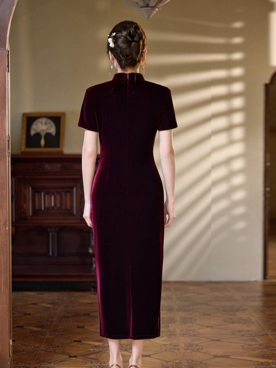 A woman viewed from behind wearing a short-sleeved, high-neck, floor-length velvet dress in deep burgundy, standing in a warmly lit indoor room with wooden floors and furniture.