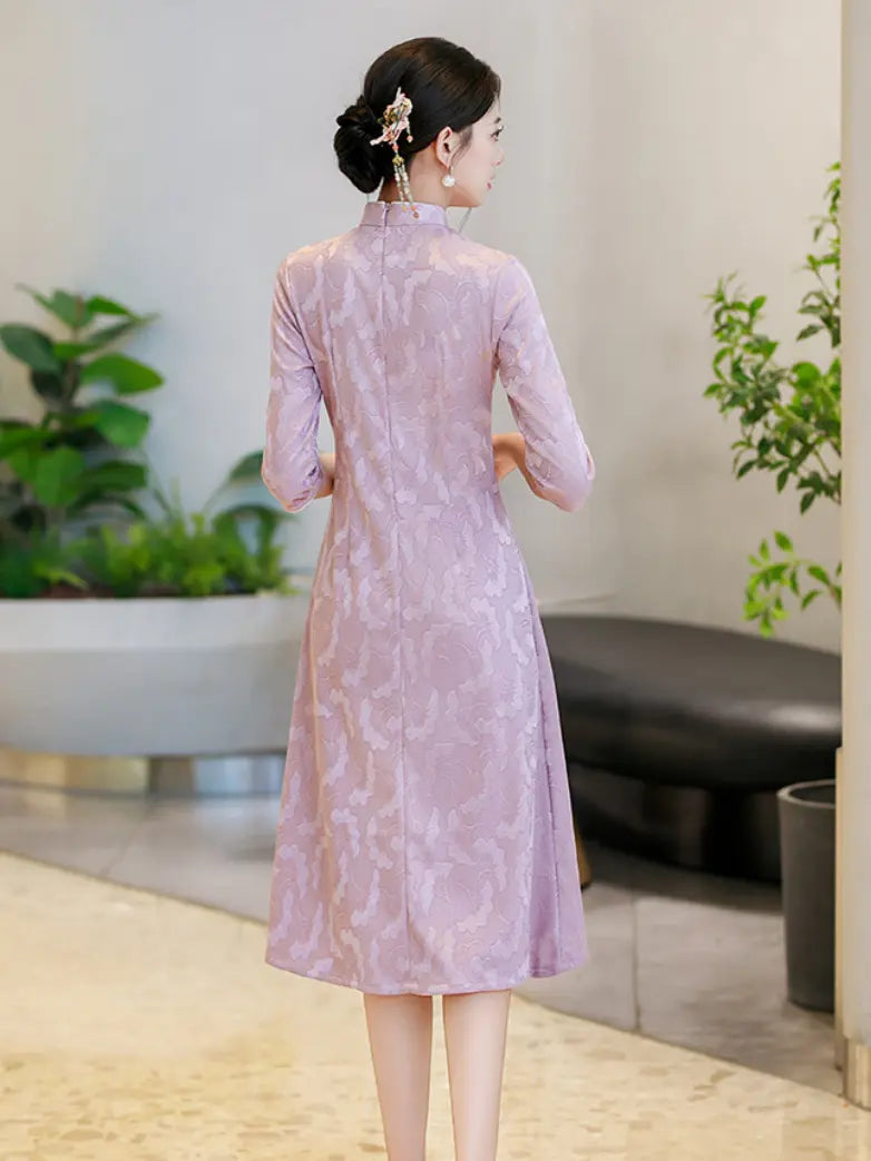A woman stands with her back to the camera wearing a lavender cheongsam dress with a subtle tonal floral jacquard pattern, set in a modern indoor environment with plants and a bench.