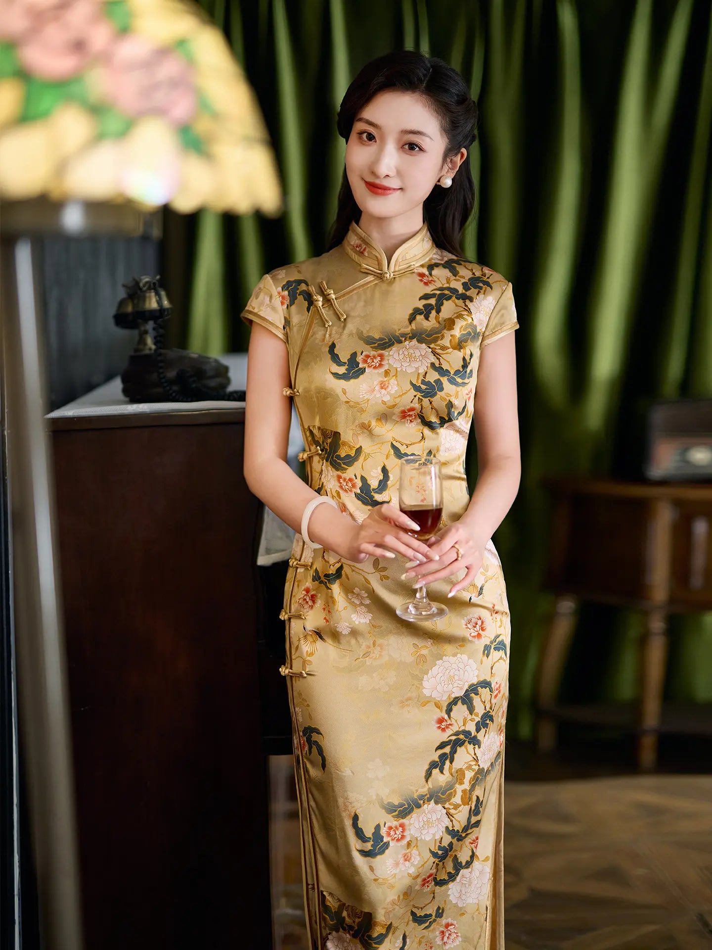 Woman in a traditional Vintage Gold Peony Silk Long Qipao Dress holding a glass in an elegant indoor setting.