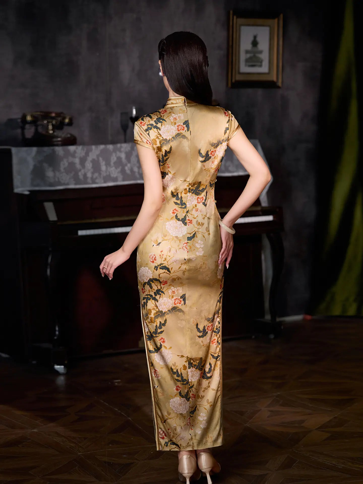 A woman viewed from behind wearing a floor-length Yellow silk cheongsam with a floral print, standing in a dimly lit room with a piano and dark wall.