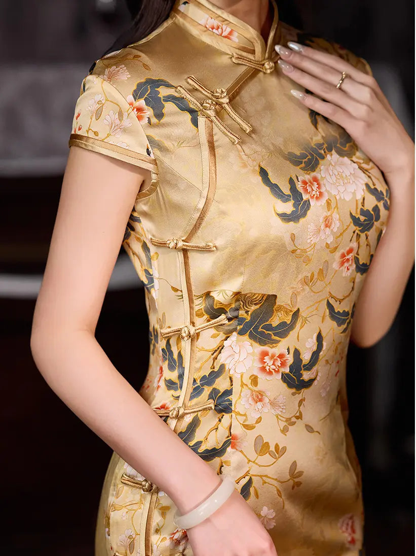 A woman wearing a short-sleeved golden yellow silk cheongsam dress with a floral pattern in pink, orange, and dark green, featuring gold-colored frog closures.