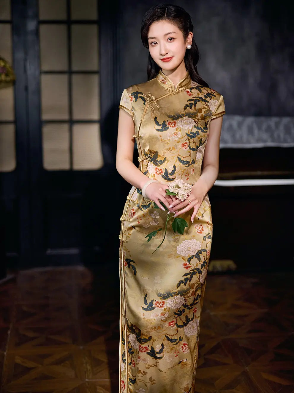 A woman wearing a Vintage Gold Peony Silk Long Qipao cheongsam Dress   with colorful floral and bird embroidery, holding a small bouquet in a dimly lit indoor setting.