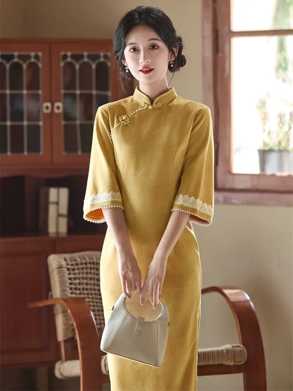 A woman wearing a yellow Chinese cheongsam (qipao) dress with lace trim on the cuffs and a decorative brooch at the collar, holding a light gray handbag in a warm, vintage-style room.