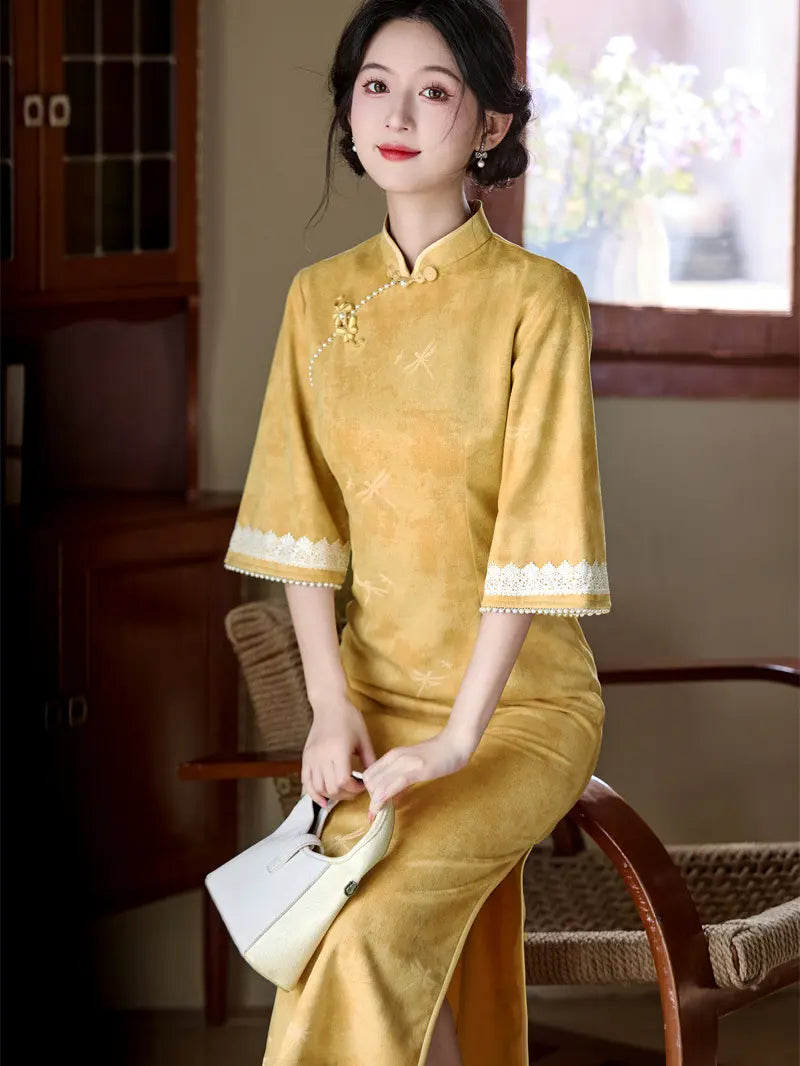 A woman wearing a yellow traditional Chinese cheongsam dress with white lace trim on the cuffs and collar, holding a small white handbag while sitting on a woven chair in a warm indoor setting.