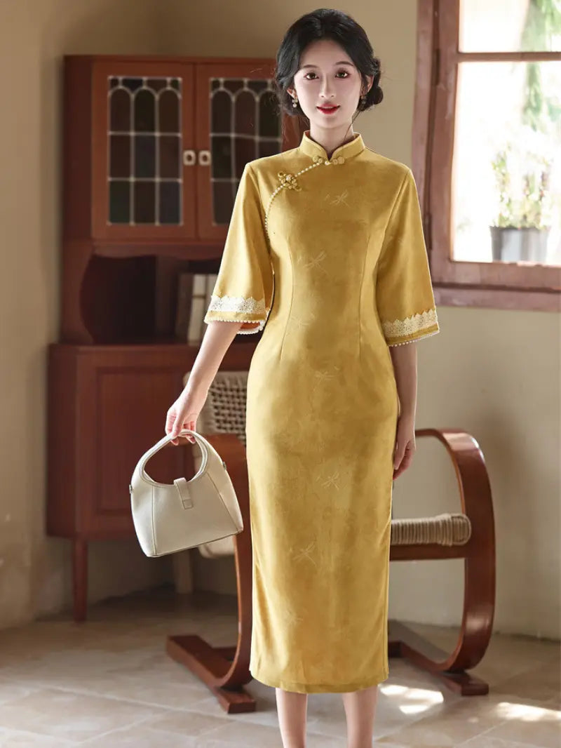 A woman wearing a mustard yellow Chinese cheongsam dress with lace trim on the sleeves, holding a small cream-colored handbag in a vintage-style room with wooden furniture.