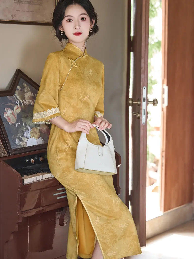 A woman wearing a mustard yellow velvet Chinese cheongsam-style dress with lace trim on the sleeves, holding a white handbag, standing in a room next to a piano and an open door.