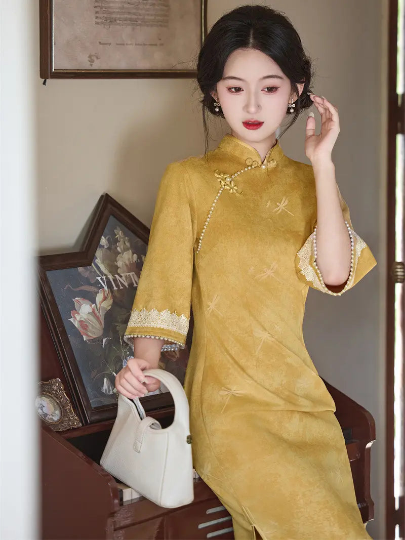 A woman wearing a mustard yellow Chinese cheongsam (qipao) dress with lace trim on the collar and cuffs, holding a white handbag in an indoor setting.