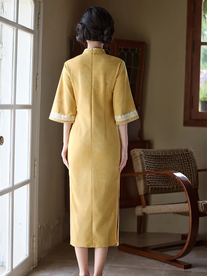 A woman viewed from behind wearing a yellow cheongsam-style dress with white lace trim at the sleeve cuffs, standing in a room with a window and wooden chair in the background.
