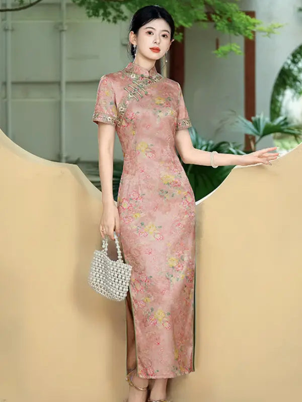 A woman models a pink floral cheongsam dress with short sleeves and a high slit, holding a small pearl handbag. She is posed against a beige sculptural backdrop with green foliage in the background.