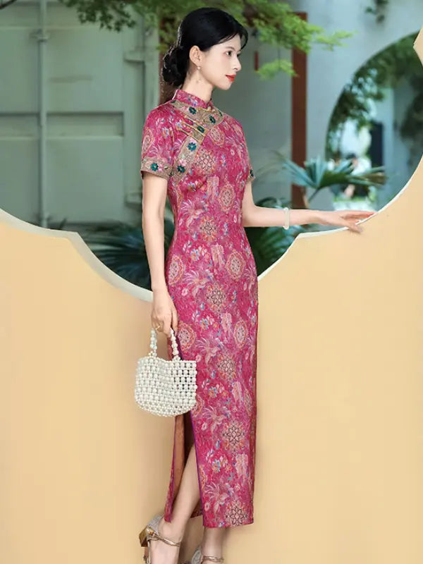 A woman wearing a pink and red floral-patterned cheongsam dress with short sleeves and a high slit, holding a white woven handbag, posing in front of a beige sculptural wall.