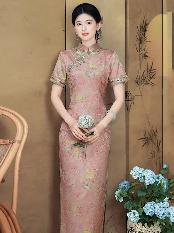 A woman wearing a pink floral cheongsam with short sleeves and a high collar, standing in front of a decorative screen. She is holding a small bouquet of white flowers, with a woven chair on the left and blue hydrangeas on the right.