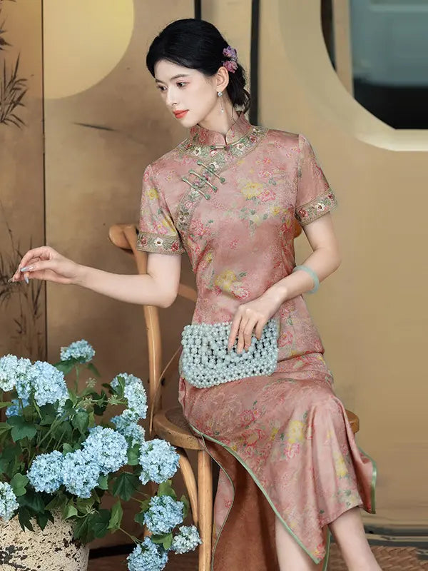 A woman seated on a wooden chair wearing a pink floral cheongsam dress with embroidered trim, holding a light blue beaded clutch. The setting includes a beige wall, decorative circular elements, and a potted plant with blue hydrangeas in the foreground.