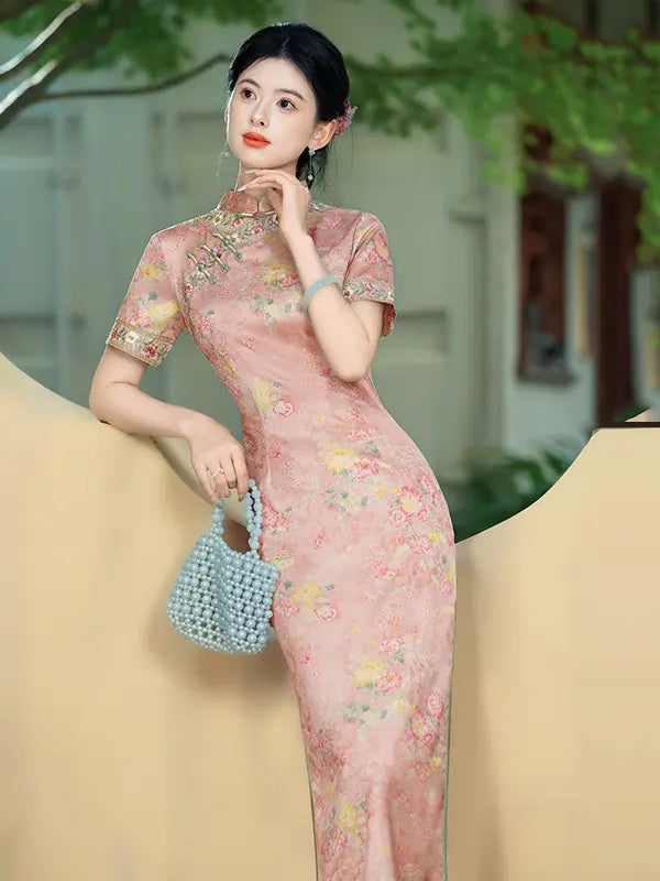 A woman wearing a pink floral cheongsam dress with short sleeves and a high collar, holding a light blue woven handbag. She is posed against a soft beige and green background.
