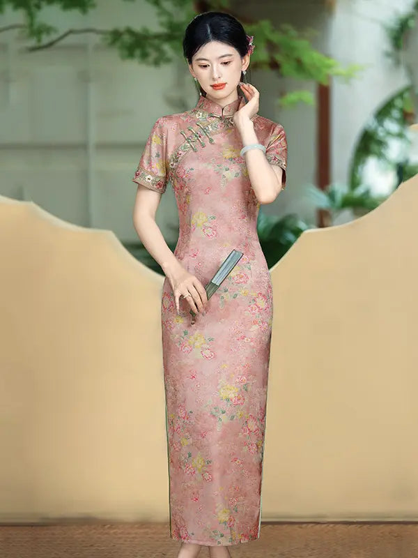 A woman wearing a pink floral cheongsam (qipao) dress with short sleeves and a high collar, standing in front of a beige background with soft green foliage.