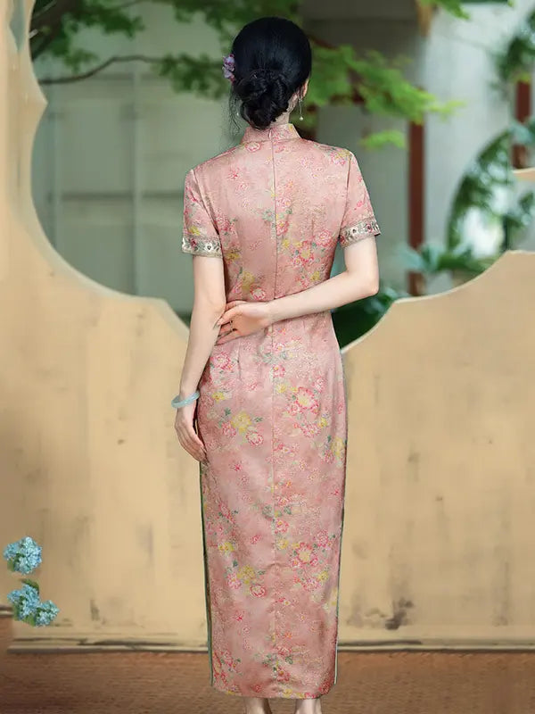 A woman viewed from behind wearing a pink floral cheongsam dress with short sleeves and lace trim at the cuffs, standing in an outdoor courtyard with a beige wall and greenery in the background.