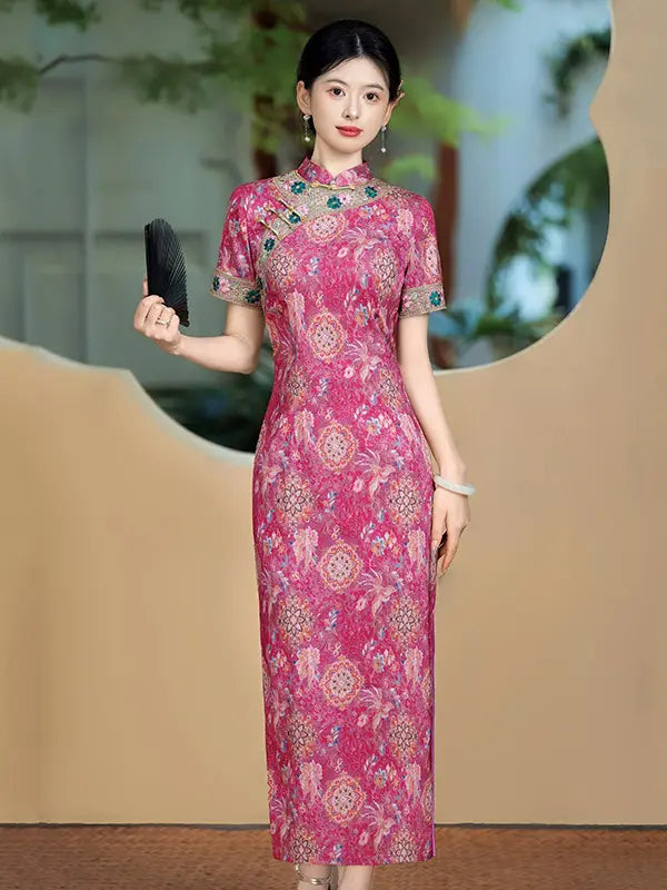 A woman modeling a pink floral-patterned cheongsam dress with green embroidered details on the collar and sleeves, holding a black folding fan against a beige background with a decorative mirror.