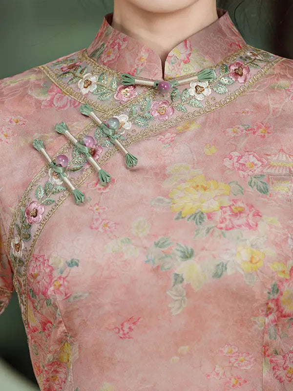 A close-up of a pink floral embroidered cheongsam-style top with green and pink decorative trim and matching decorative buttons, worn by a model.