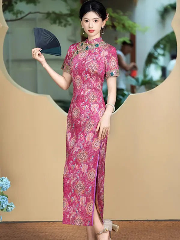 A woman models a pink floral-patterned cheongsam dress with short sleeves and a high slit, holding a black folding fan. The background features a beige decorative wall with a circular cutout showing a softly blurred outdoor scene.