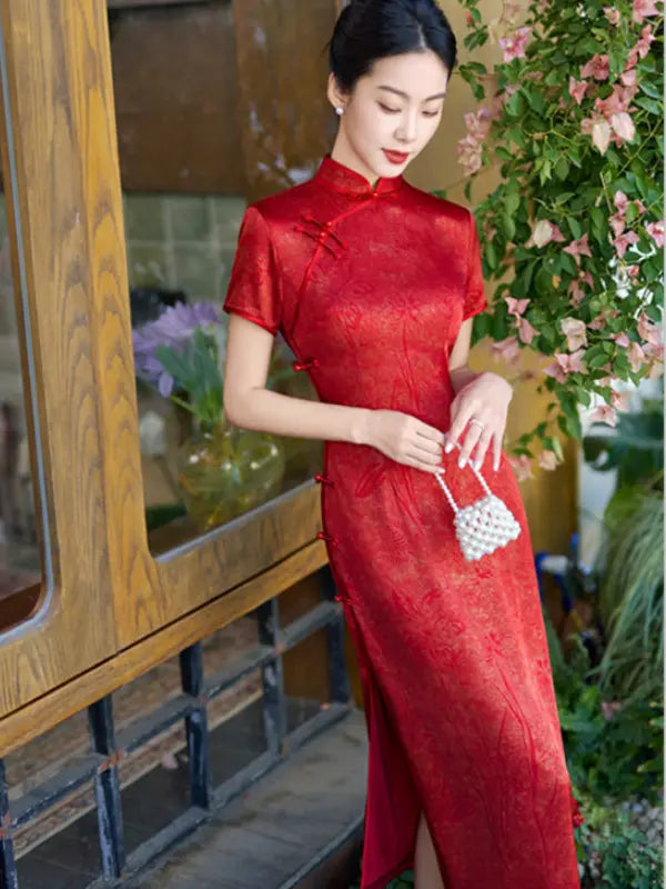 A woman wearing a short-sleeved red cheongsam/qipao dress with a subtle floral jacquard pattern, standing in front of a wooden door with green foliage and pink flowers in the background.