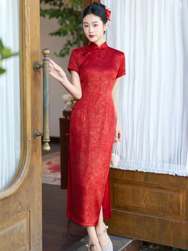 A woman wearing a red short-sleeve cheongsam/qipao dress with a subtle tonal floral pattern, standing in a doorway next to a wooden door handle with white curtains in the background.