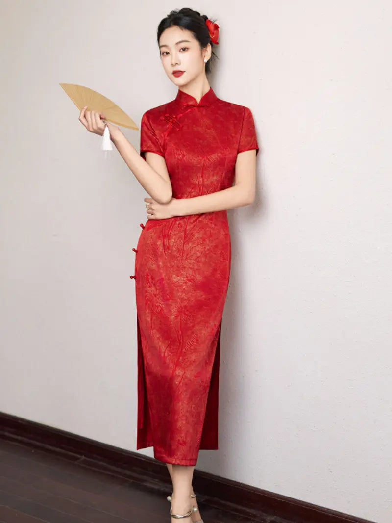 A woman wearing a red short-sleeve cheongsam dress with a subtle tonal floral pattern, holding a beige folding fan against a plain off-white wall.