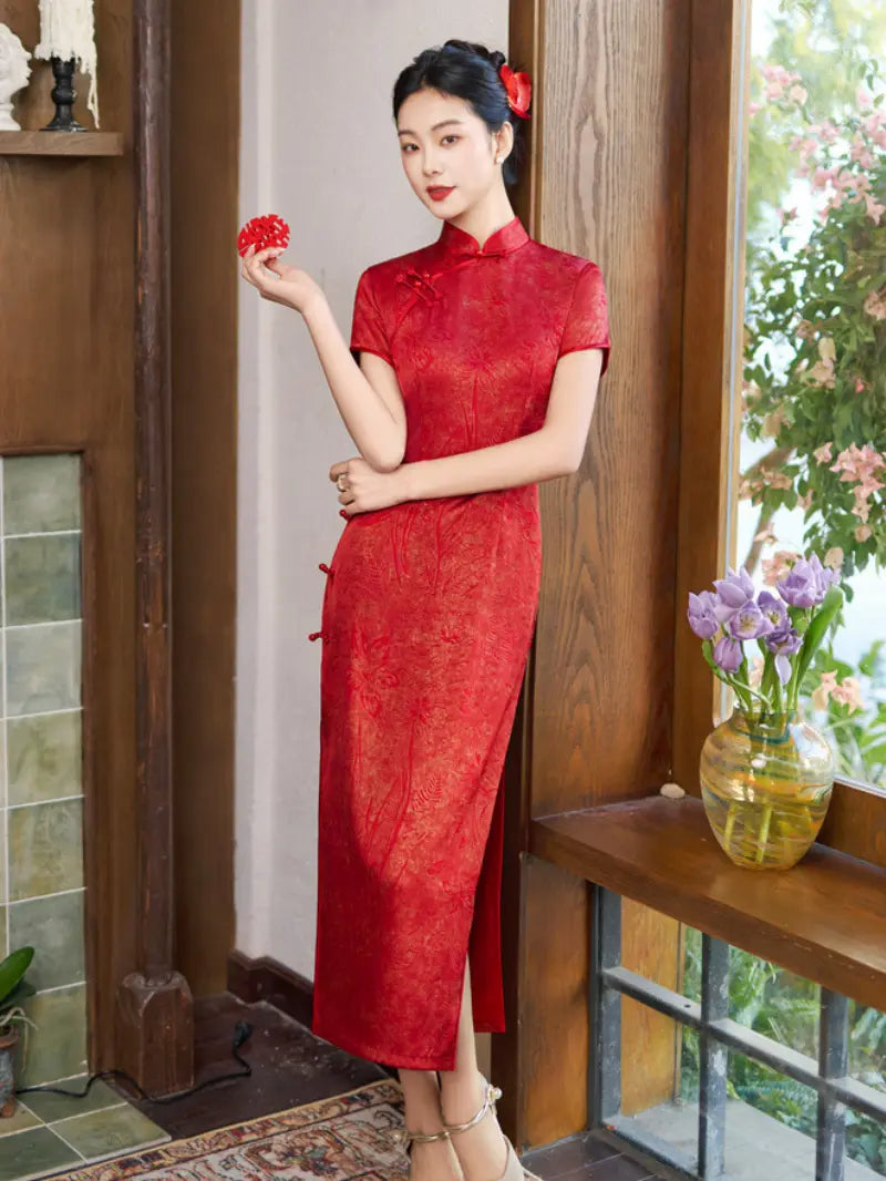 A woman wearing a red short-sleeve cheongsam/qipao dress stands indoors near a wooden window and shelf, holding a small red flower. The dress has a subtle tonal pattern and side slit.