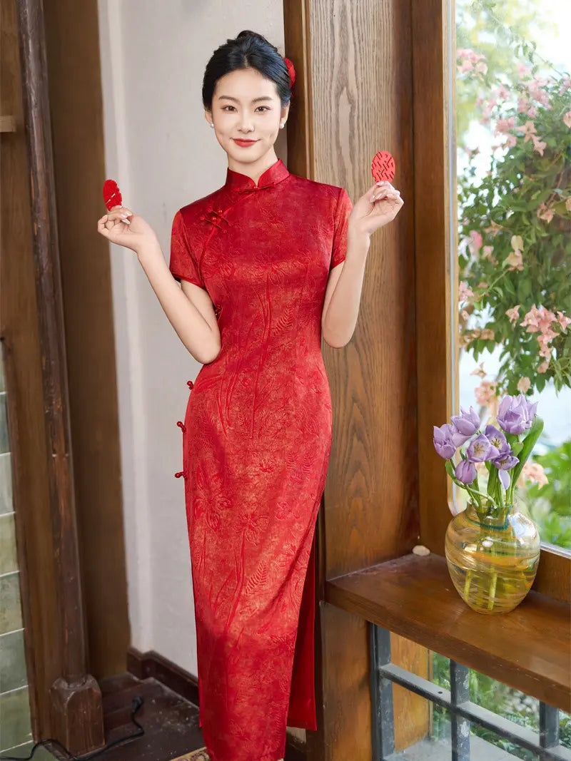 A woman wearing a red embroidered cheongsam/qipao dress stands indoors near a wooden window frame, holding two red decorative items, with flowers in a glass vase on the windowsill beside her.
