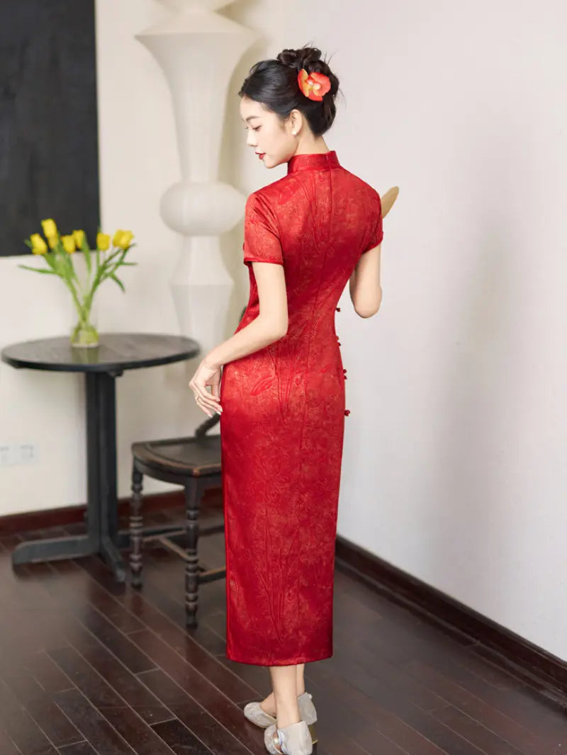 A woman wearing a red short-sleeved cheongsam dress, photographed from behind in an indoor setting with a dark wood floor, white wall, and a small table with yellow tulips.