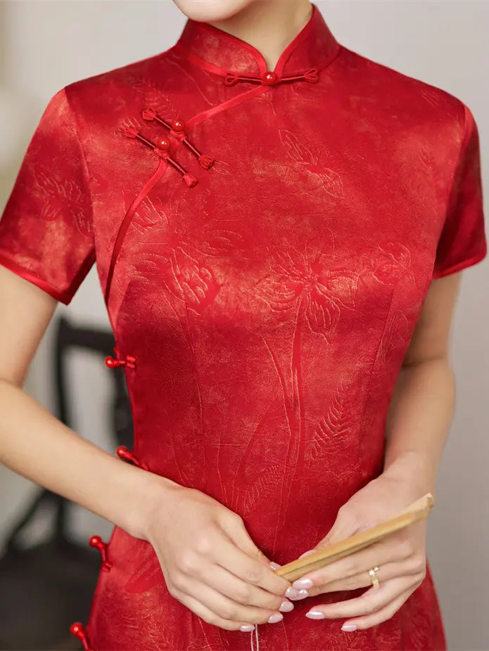 A close-up of a person wearing a short-sleeve red cheongsam/qipao dress with tonal floral jacquard texture and red button-and-chinstrap details.