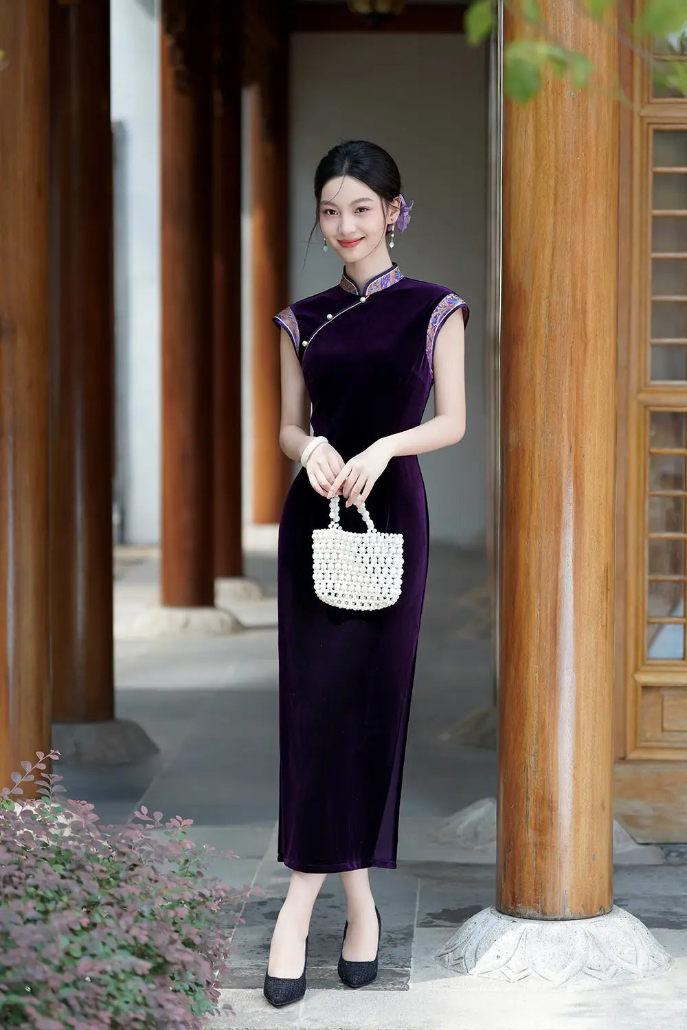 Woman in a Violet Velvet Qipao Cheongsam Dress with Floral Trim holding a white handbag in an outdoor setting.