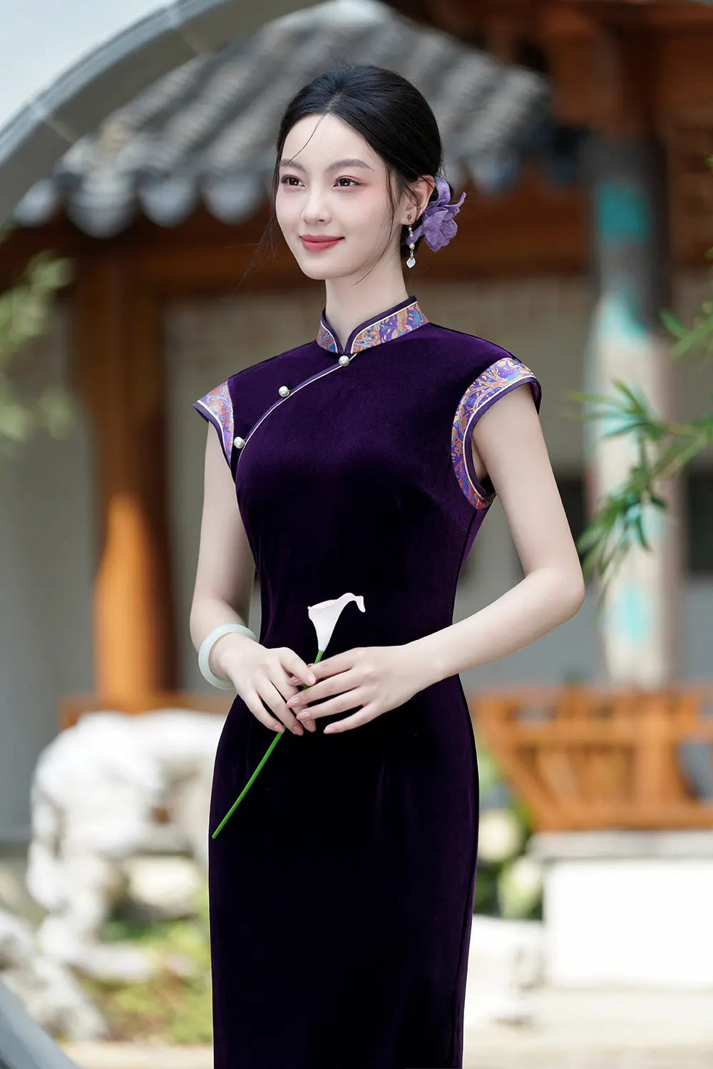 Woman in a traditional Chinese Violet Velvet Qipao Cheongsam Dress with Floral Trim standing outdoors with a blurred background