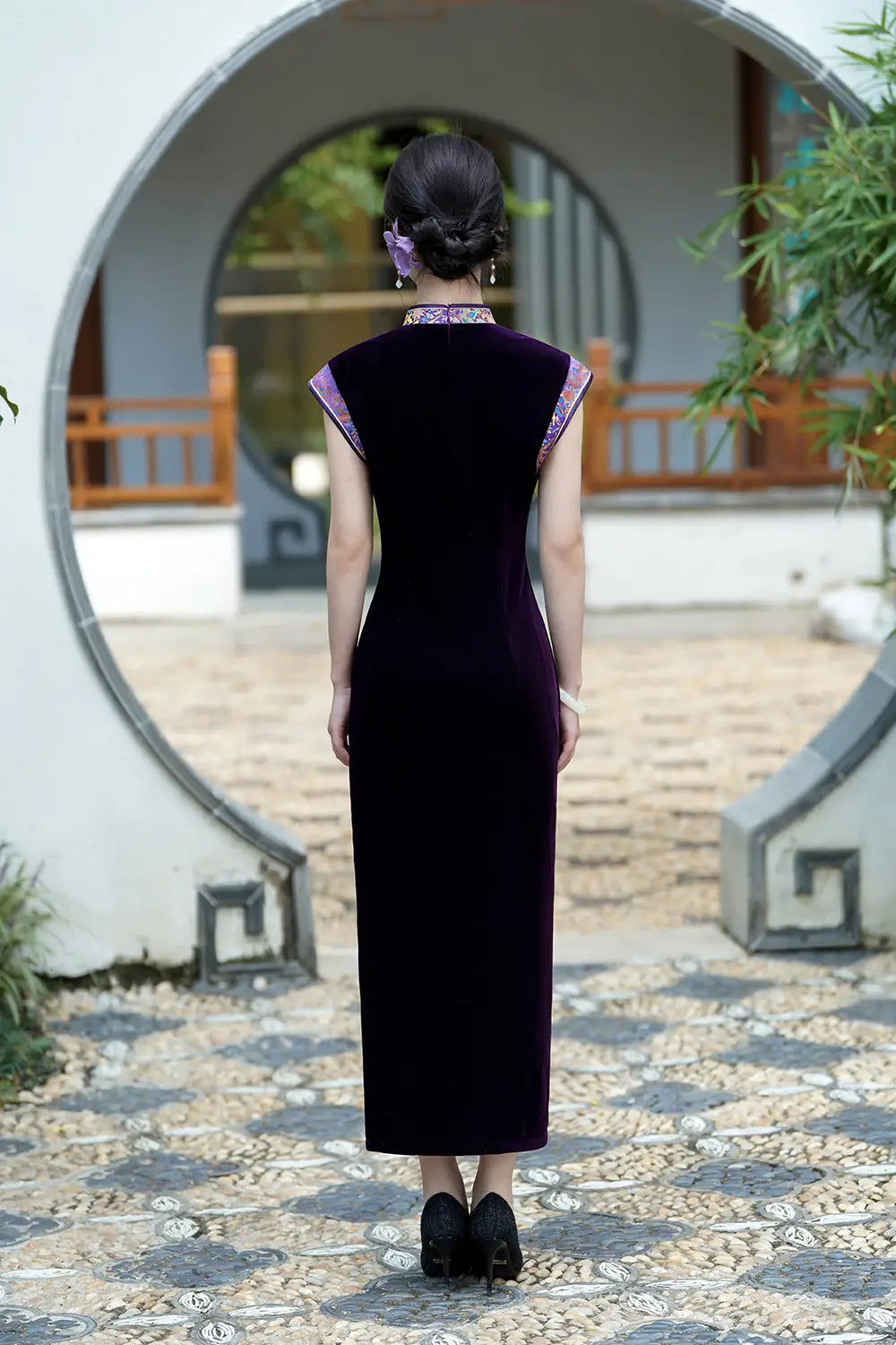 Woman wearing a long Violet Velvet Qipao Cheongsam Dress with Floral Trim  standing in front of a traditional Chinese garden gate.