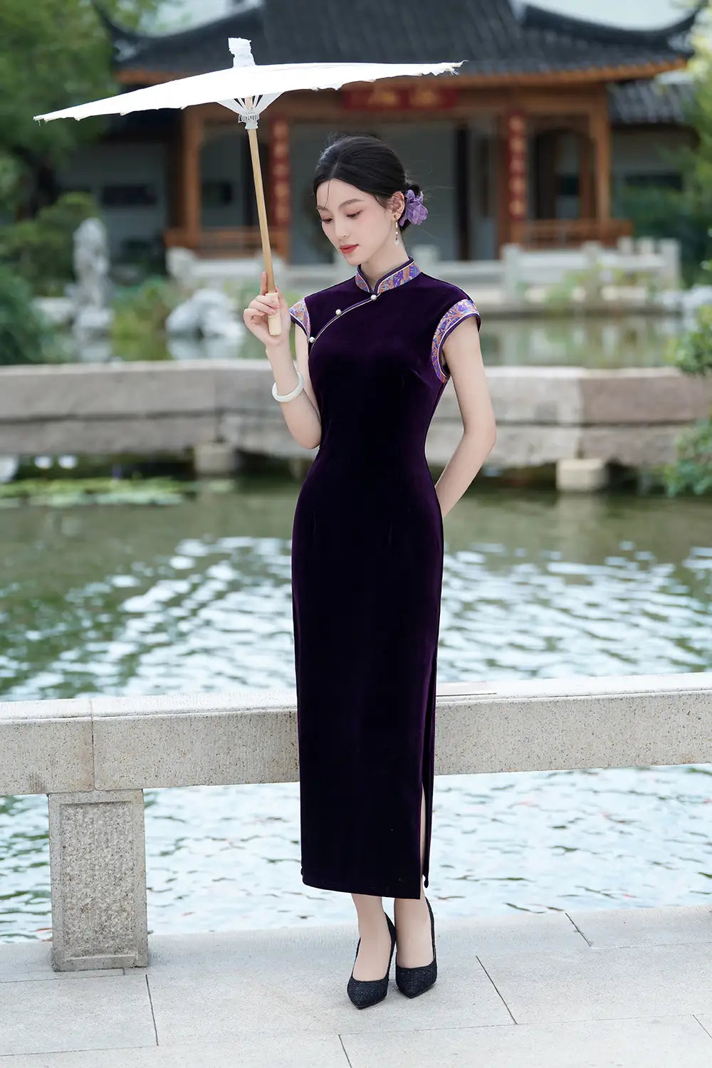Woman in a long Violet Velvet Qipao Cheongsam Dress with Floral Trim holding an umbrella in a garden setting