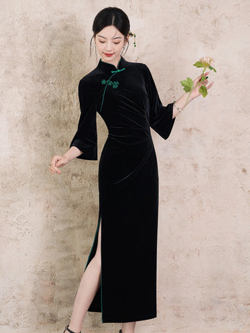 A woman models a black velvet long sleeve qipao cheongsam dress with a high side slit and green embroidery at the collar, standing against a textured beige wall and holding a small flower.