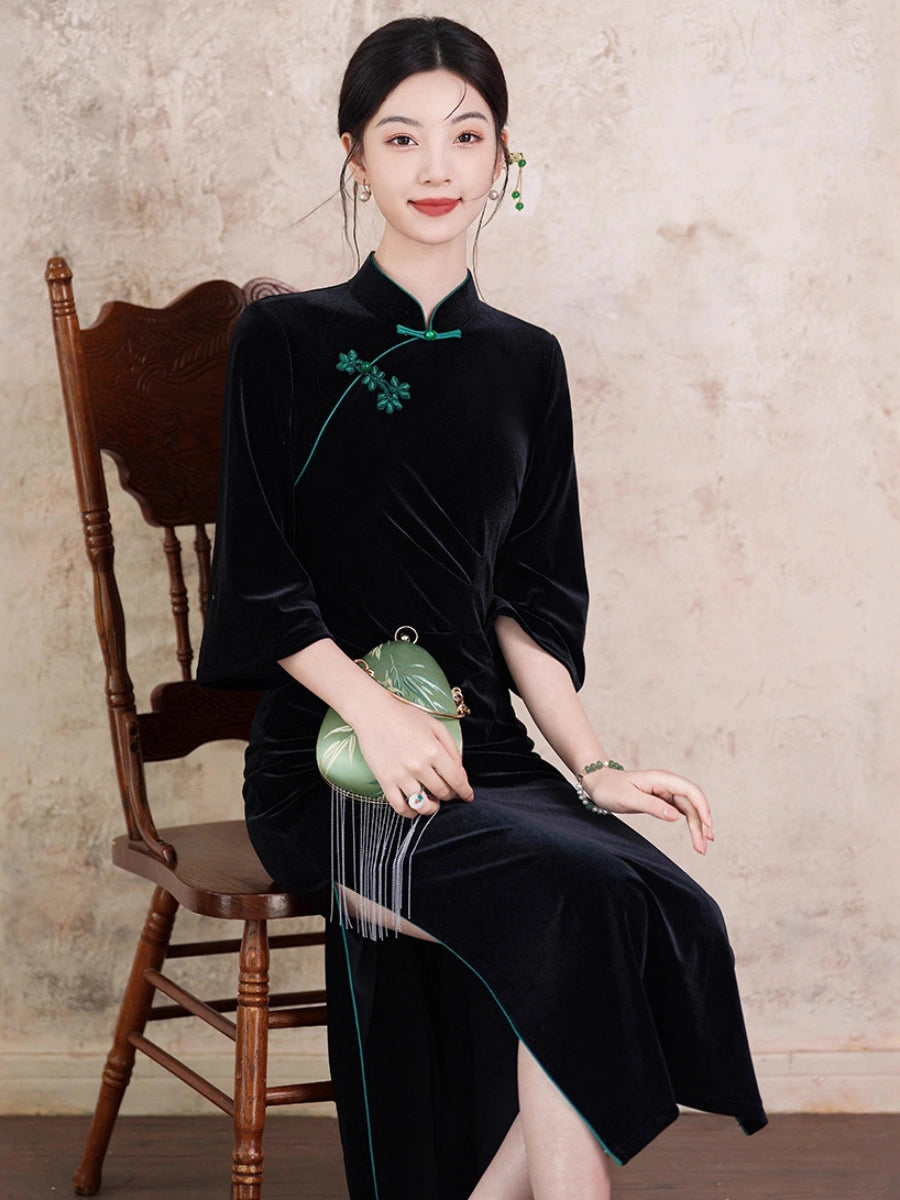 A woman sits on a wooden chair wearing a black velvet cheongsam-style dress with green embroidery and a green bow at the collar. She holds a small green handbag against a textured beige wall.