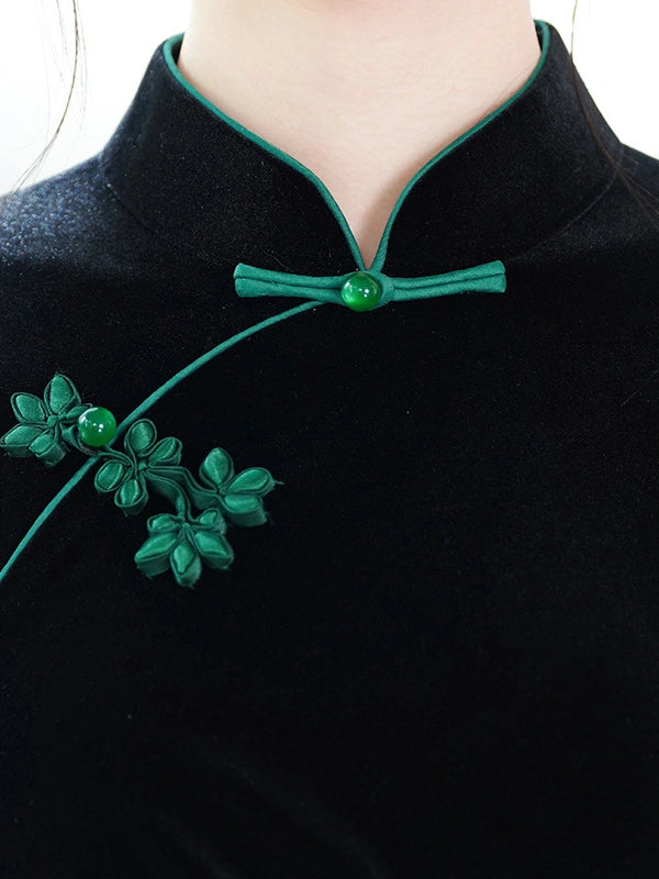 Close-up of a black velvet traditional Chinese-style top with green cord trim, a green bow closure, and a green floral brooch on the chest.