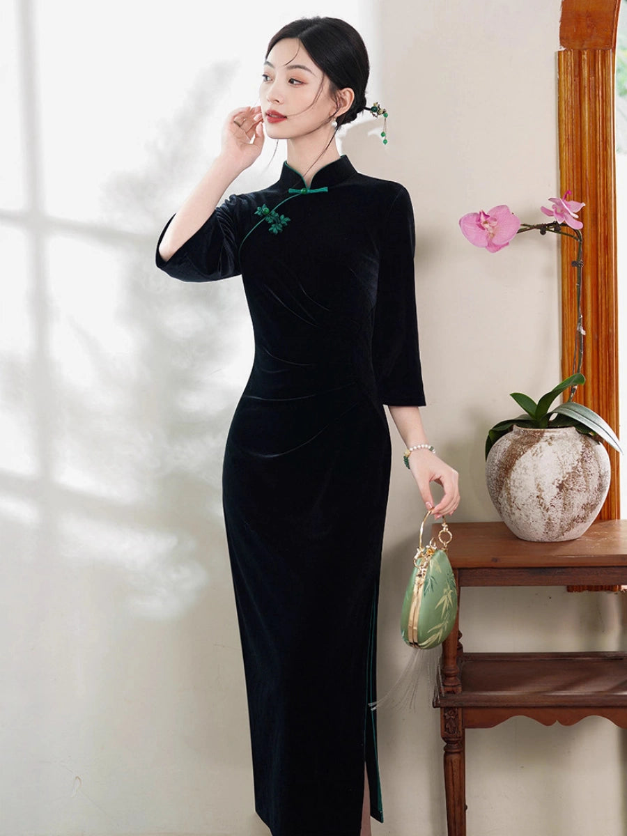 A woman models a black velvet cheongsam dress with green embroidery and a green collar, standing in a styled indoor setting next to a wooden table with a vase of pink flowers.
