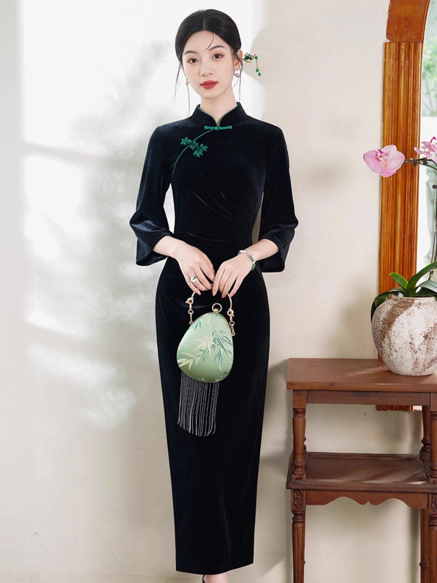 A woman stands indoors wearing a black velvet qipao  cheongsam-style dress with green trim and a green floral chest detail, holding a light green embroidered handbag. The setting includes a cream wall, a wooden side table, and a vase with pink flowers.