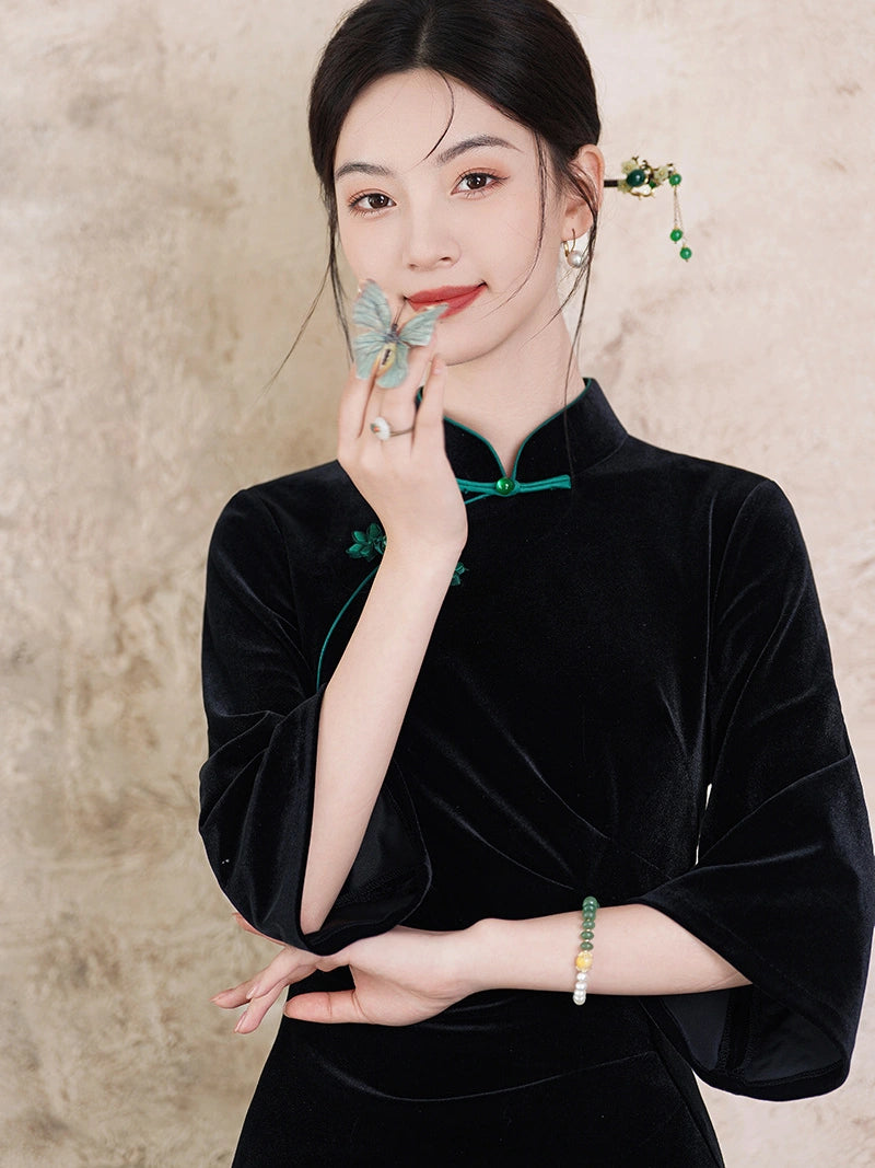 A woman wearing a black velvet cheongsam-style top with green trim, posed against a textured beige wall. She is holding a butterfly ornament near her face and wearing green and white beaded jewelry.