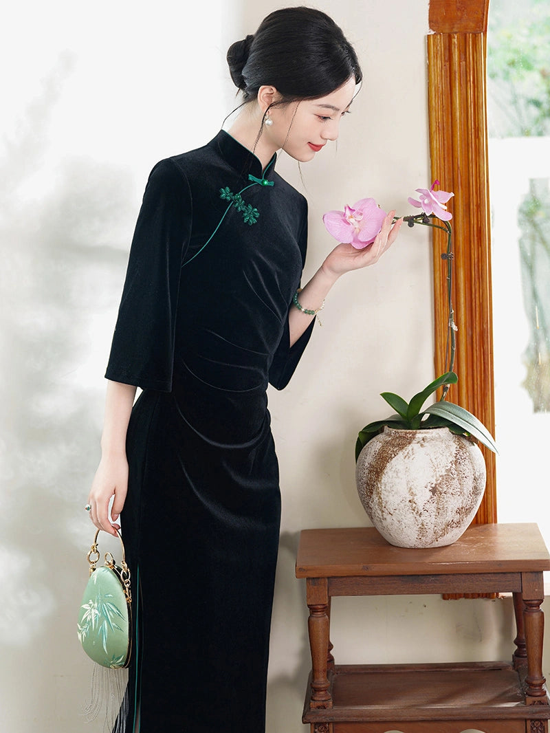 A woman wearing a black velvet cheongsam dress with green embroidery stands beside a wooden table holding a pink orchid, holding a light green embroidered handbag.