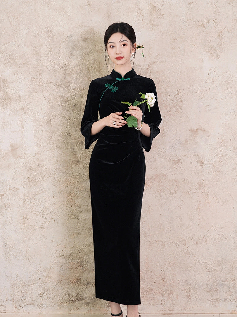 A woman stands against a textured beige wall wearing a long black velvet cheongsam-style dress with green embroidery and a green cord at the collar, holding a white flower with green leaves.