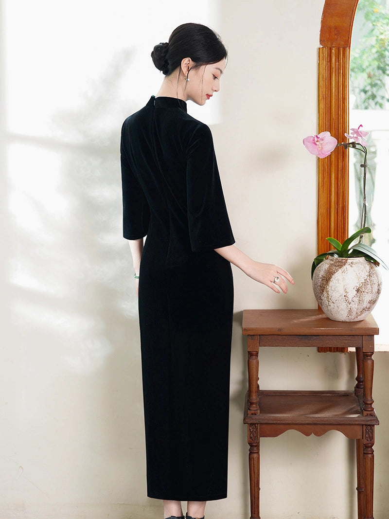 A woman wearing a black velvet three-quarter sleeve cheongsam-style dress stands in profile beside a small wooden table with a potted pink orchid against a cream wall.