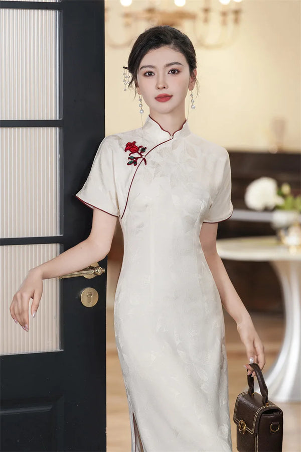 Woman in a white traditional Jacquard Qipao Cheongsam Dress With Red Trimwith red floral embroidery, holding a brown handbag indoors.