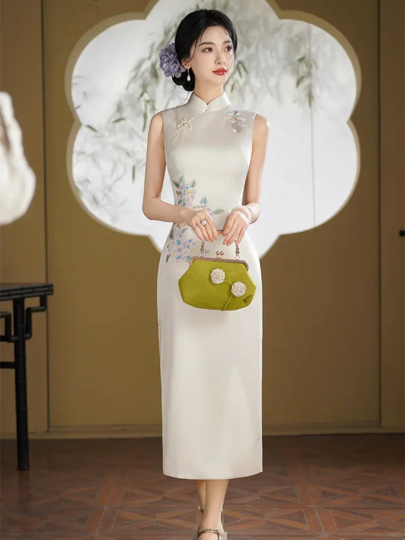 A woman models a sleeveless cream-colored Chinese cheongsam dress with floral embroidery on the skirt, holding a green handbag in a traditional indoor setting.