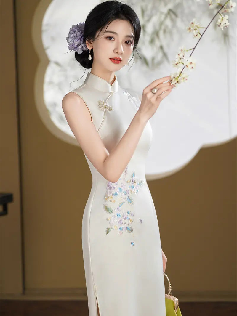 A woman wearing a sleeveless cream-colored Chinese cheongsam dress with delicate floral embroidery on the skirt, holding a flowering branch in a styled indoor setting.