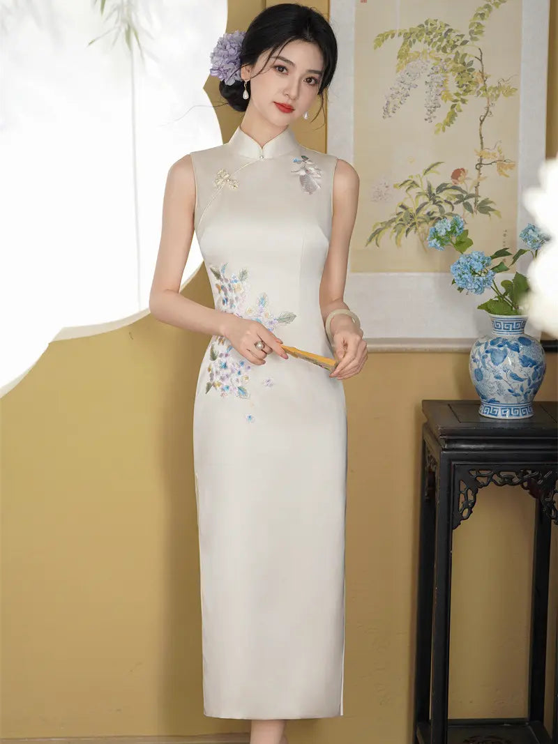 A woman models a sleeveless cream-colored Chinese cheongsam dress with delicate blue and green floral embroidery on the bodice and hips. She is standing in a decorated room with a yellow wall, a traditional painting, and a blue-and-white ceramic vase on a dark wooden table.
