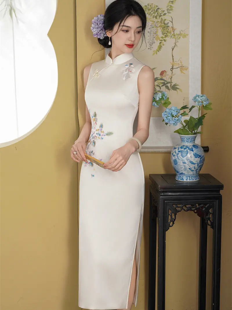 A woman wearing a sleeveless cream-colored cheongsam with delicate floral embroidery stands in a traditional room setting with a yellow wall, a black wooden side table, and a blue-and-white ceramic vase.