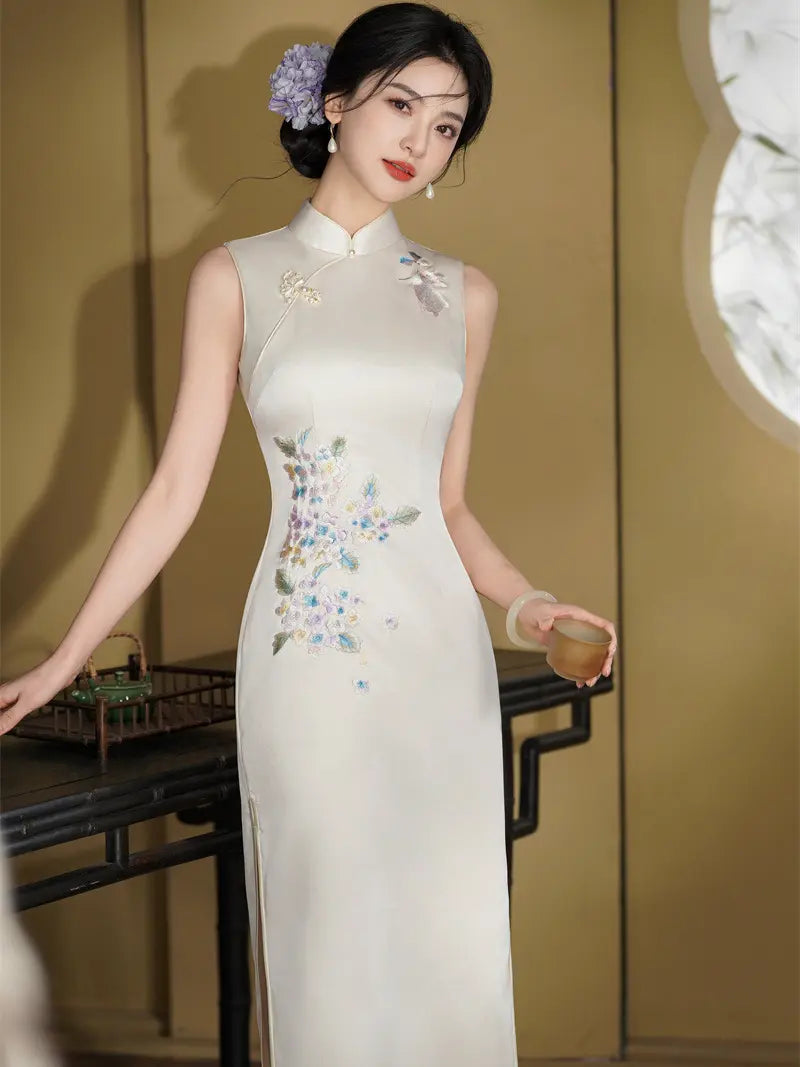 A woman wearing a sleeveless cream-colored Chinese cheongsam dress with floral embroidery on the bodice and skirt, standing in a warm-toned indoor setting.