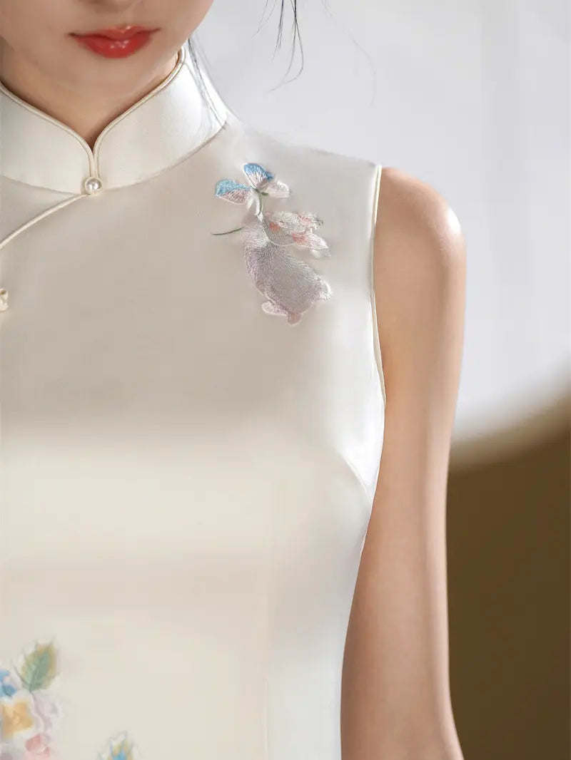 Close-up of a woman wearing a sleeveless cream-colored cheongsam-style dress with embroidered floral details on the chest and shoulder, against a softly blurred neutral background.